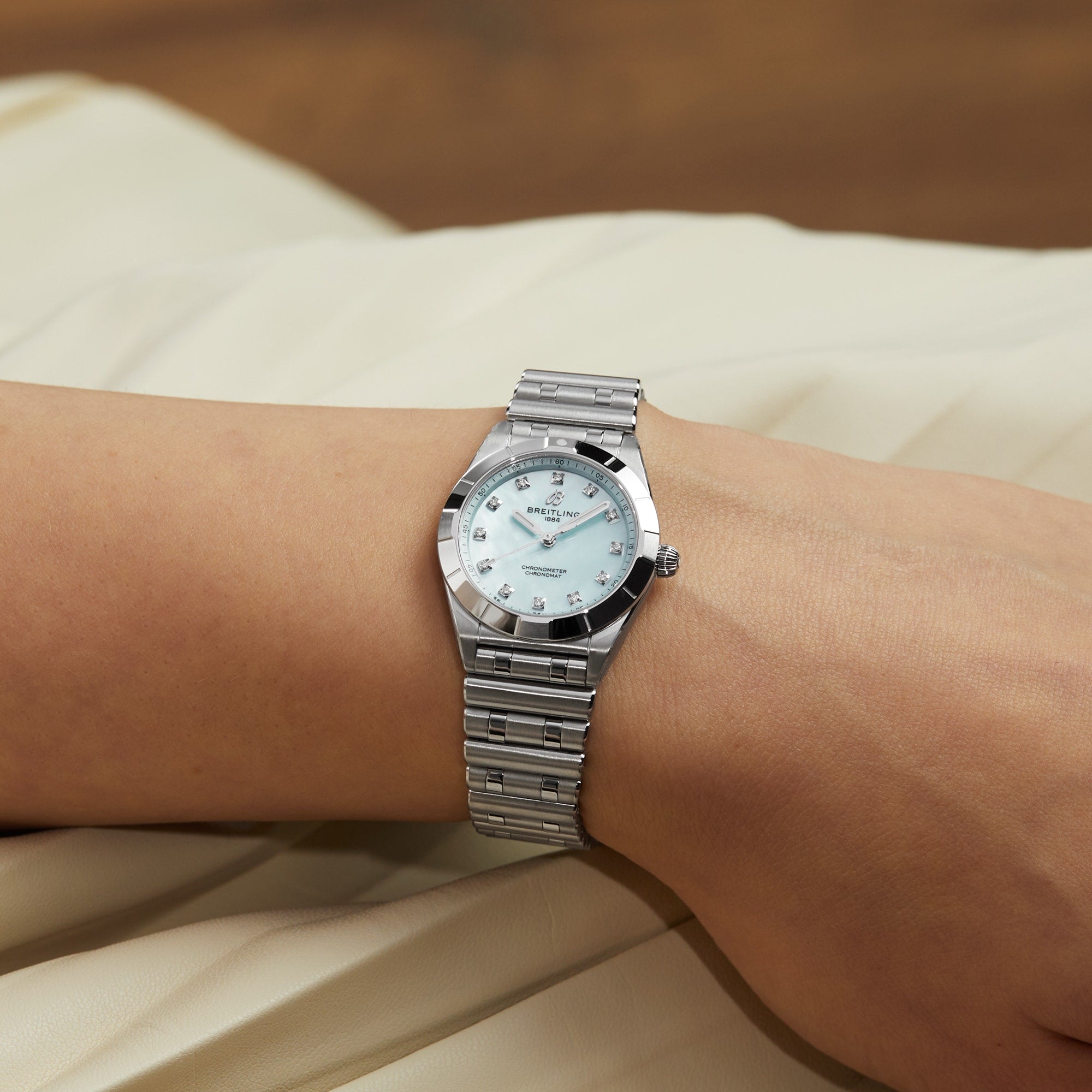 A Breitling Chronomat 28 wristwatch with a silver metal band and light dial is worn on a person’s wrist, resting on a pale fabric surface.
