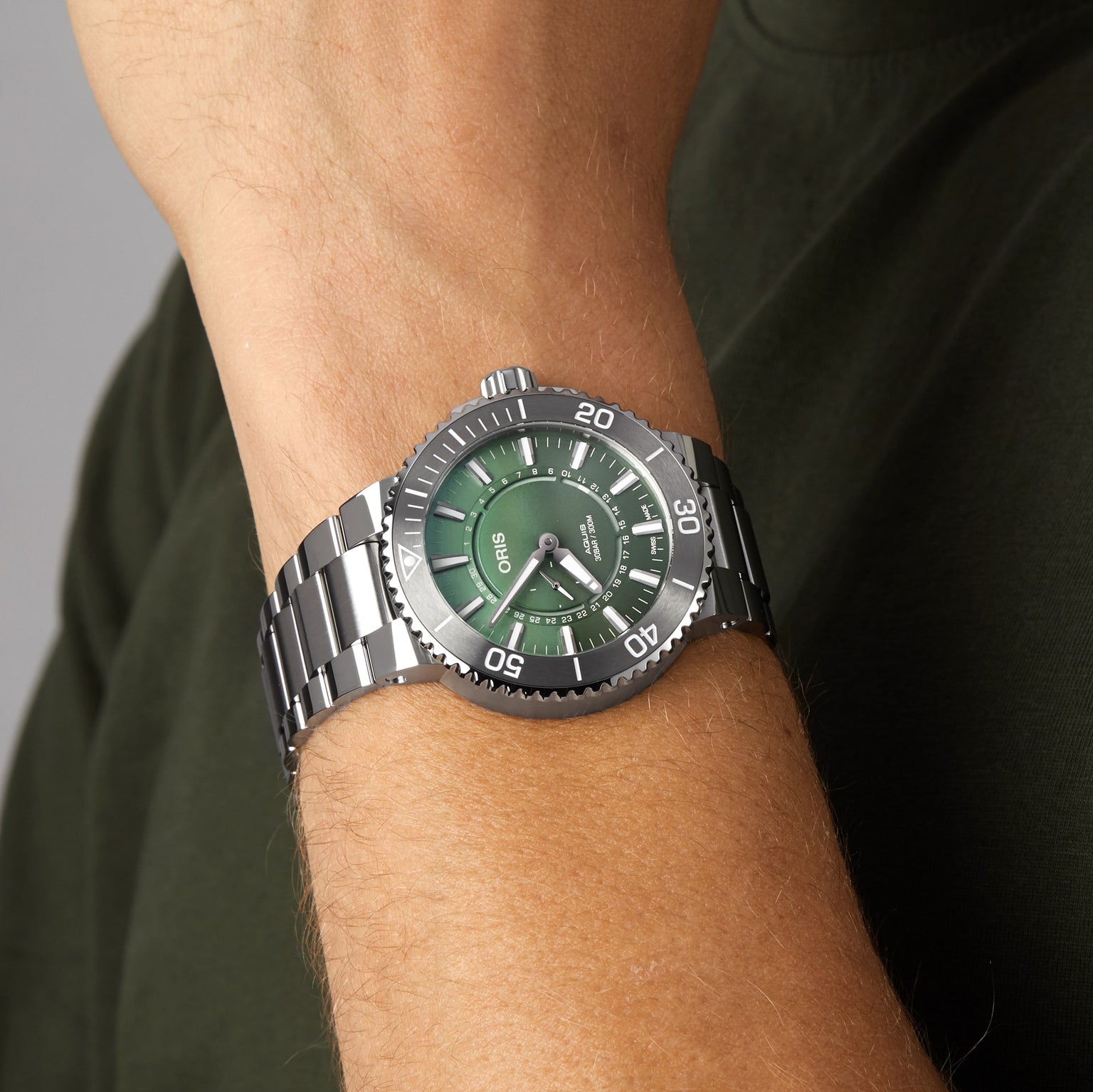 An Oris Dat Watt Limited Edition II silver wristwatch with a green dial is worn on a person’s wrist, paired with a dark green shirt.