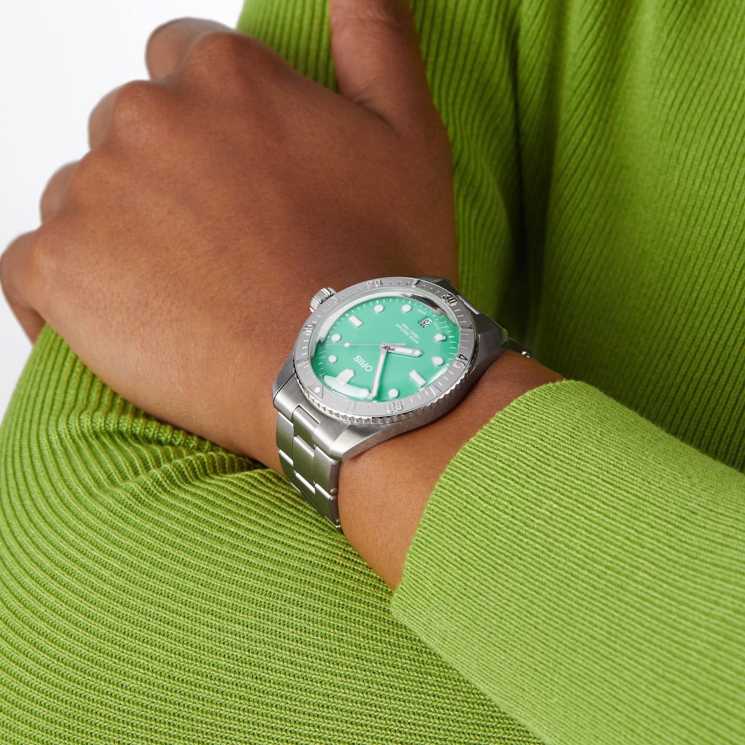 A person wears the Oris Divers Sixty‑Five Cotton Candy silver watch with a green face, paired with a bright green ribbed long-sleeve top.