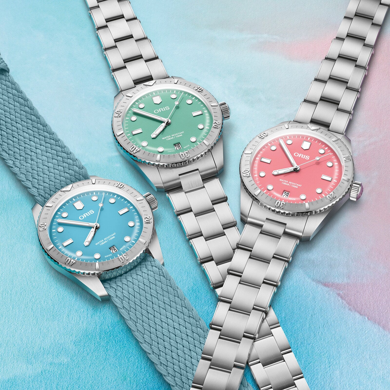 Three Oris Divers Sixty‑Five Cotton Candy watches—with blue, green, and pink dials—are displayed on a pastel background; two feature metal straps, while one has a fabric strap.