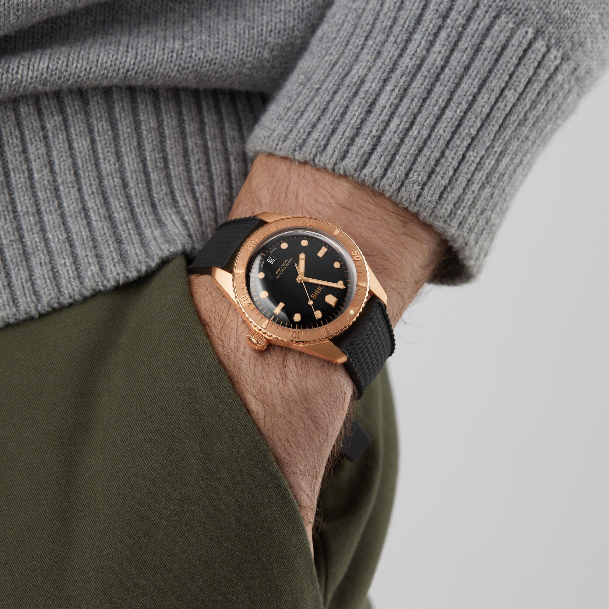 A person wearing the Oris Divers Sixty‑Five Date gold watch with a black strap and a grey sweater, hand in olive green pants pocket.
