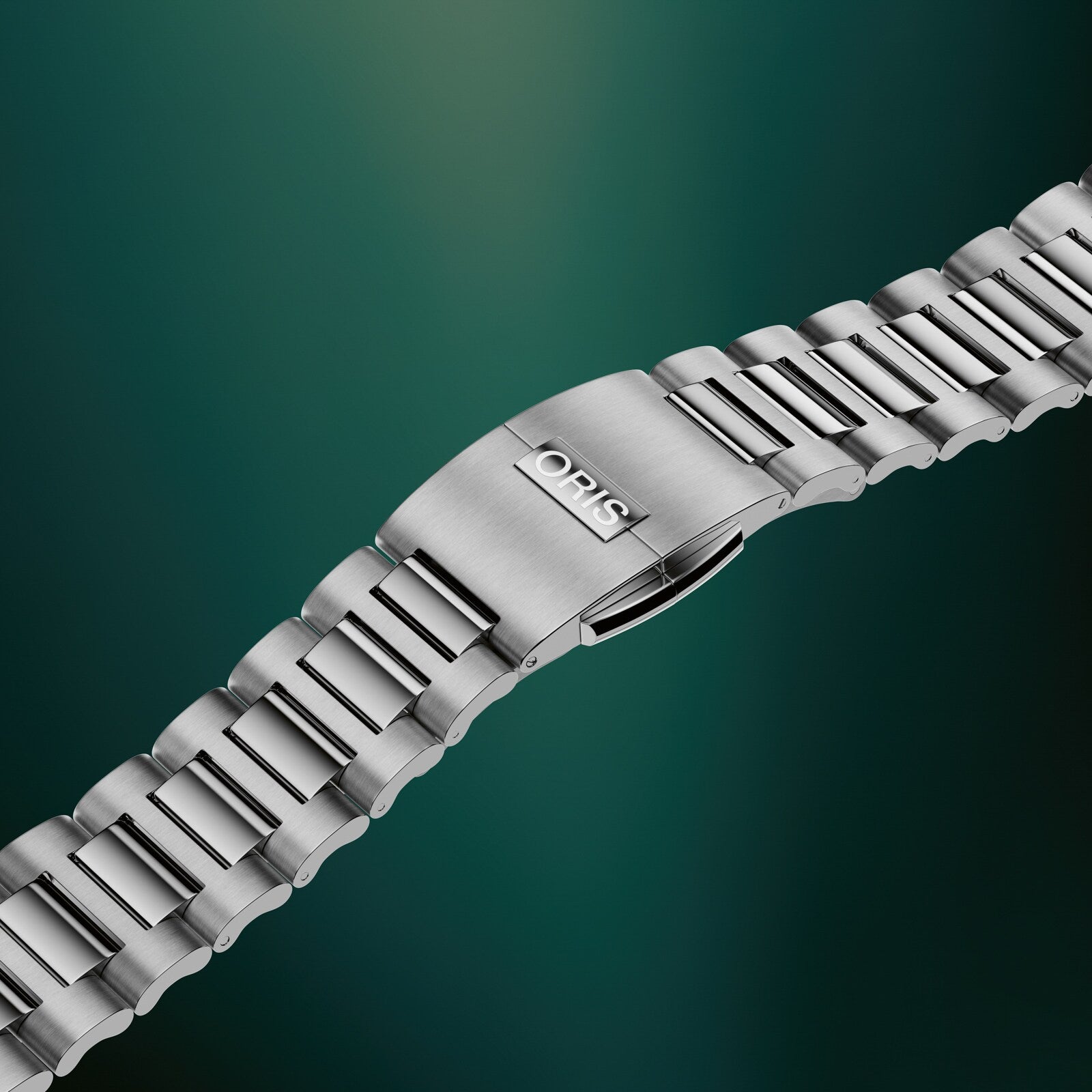 Silver-tone metal watch bracelet with link construction and fold-over clasp against teal background.