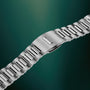 Silver-tone metal watch bracelet with link construction and fold-over clasp against teal background.