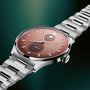 Stainless steel watch with terracotta dial, moonphase display, subdial, and matching link bracelet on teal background.
