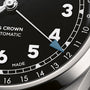 Watch face with black dial, white Arabic numerals, blue pointer hand, date window, and tachymeter scale on silver case.