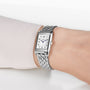 A close-up of a wrist wearing the Jaeger-LeCoultre Reverso Classic, featuring a silver rectangular case, metal link band, off-white dial with black numerals and blue hands. The diamond bezel complements a light-colored long-sleeve shirt.