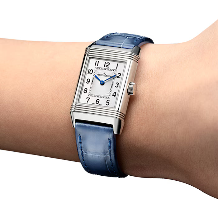 A Jaeger-LeCoultre Reverso Small Duetto rectangular watch with a white dial, black Arabic numerals, blue hands, and a blue leather strap is worn on someone's wrist.