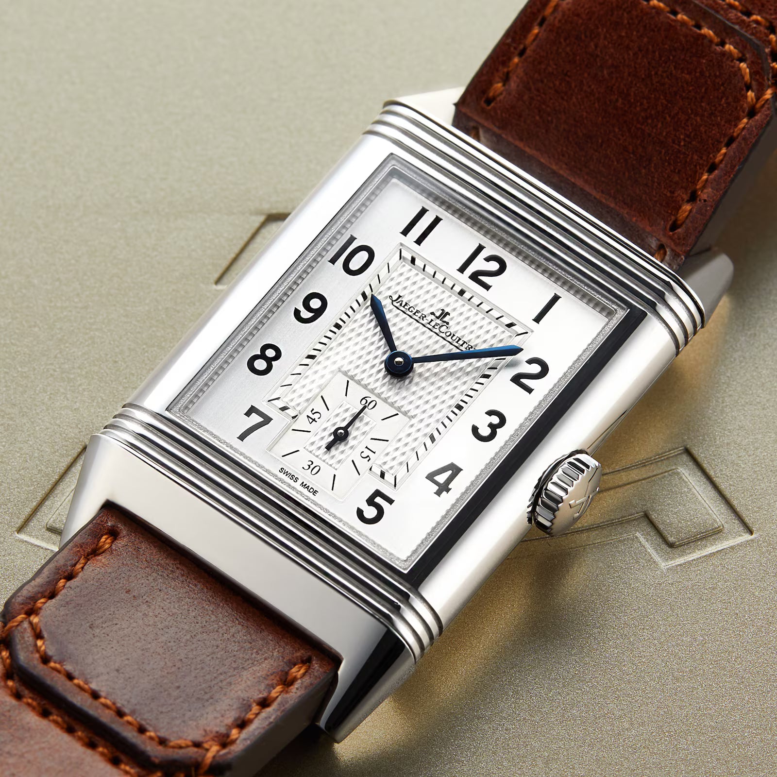 A rectangular Jaeger-LeCoultre Reverso Classic Large Small Seconds watch with a white dial, black numerals, blue hands, and a brown leather strap is displayed on an embossed metallic surface.