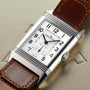 A rectangular Jaeger-LeCoultre Reverso Classic Large Small Seconds watch with a white dial, black numerals, blue hands, and a brown leather strap is displayed on an embossed metallic surface.