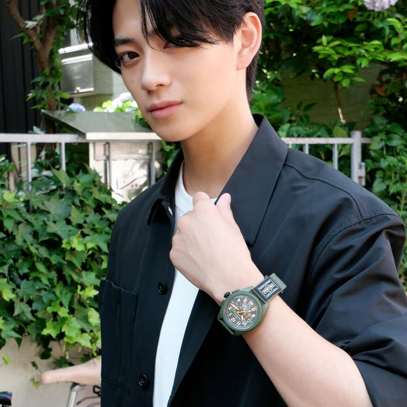 A person outdoors, in a black shirt over a white tee, wears the Zenith Limited Edition Pilot Big Date Flyback Porter on their left wrist. Lush green plants and a fence appear in the background.