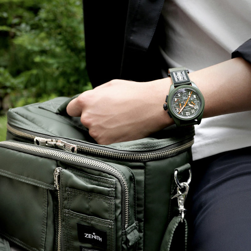 A person wearing a Limited Edition Zenith Pilot Big Date Flyback Porter rests their arm on a matching green Zenith bag outdoors.