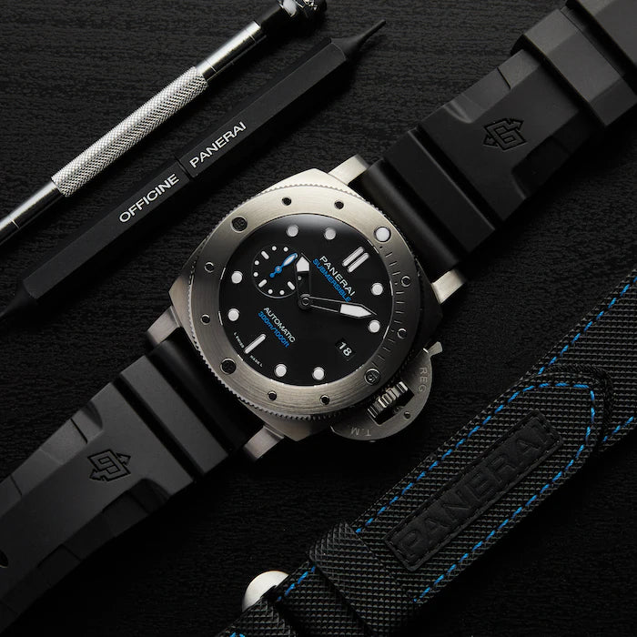 A close-up of the Panerai Submersible es men’s dive watch with a titanium case, black dial with blue accents, date display, and rubber strap, surrounded by branded watch tools on a dark surface.