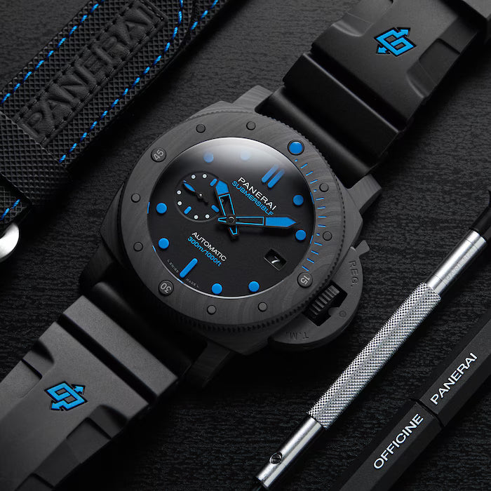 A Panerai Submersible Carbotech wristwatch with blue accents rests on a black surface beside a black strap, Panerai tool, and logo case—highlighting its robust carbon fibre build for divers.