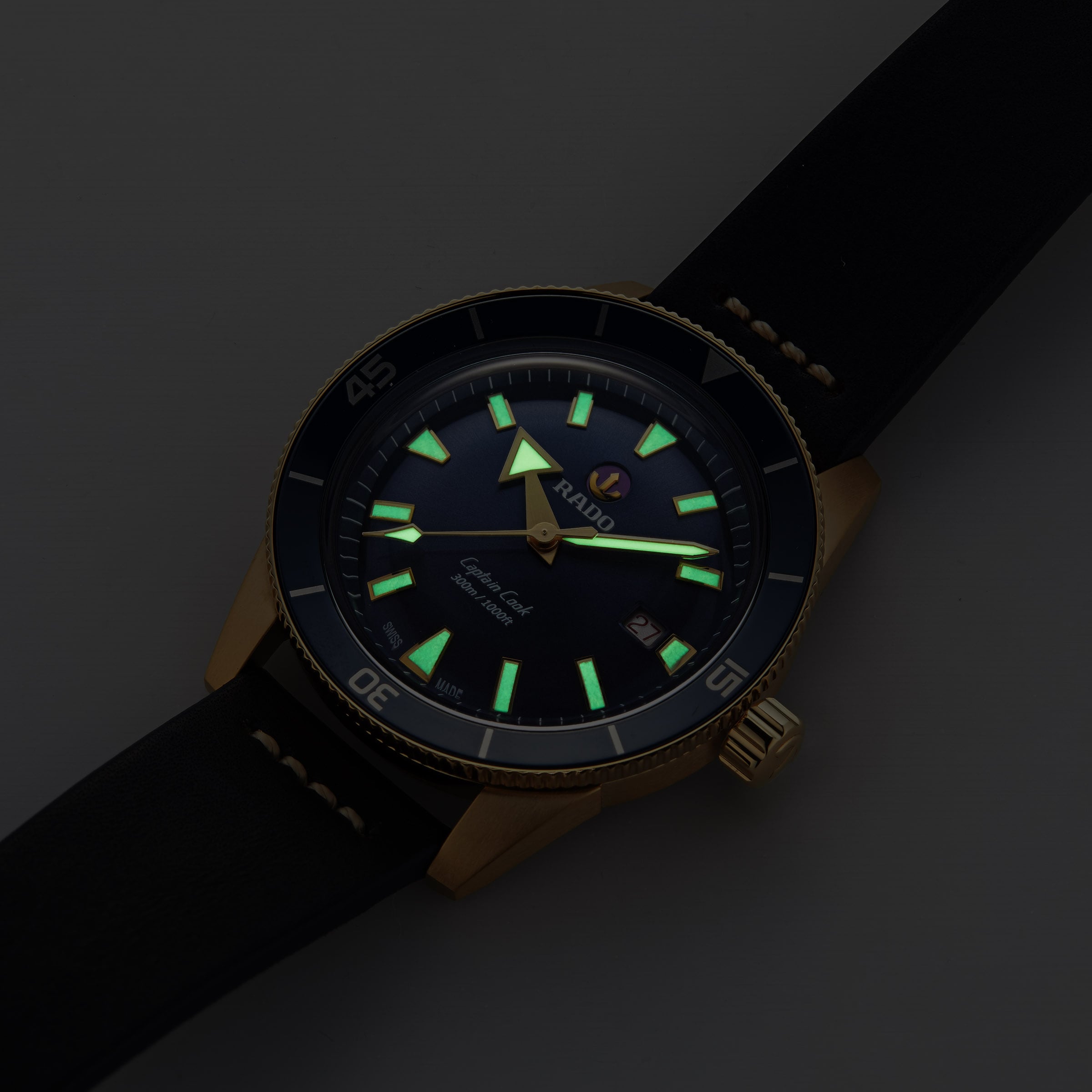 The Rado Captain Cook Automatic Bronze wristwatch features a black dial, bronze case, and black strap with luminous hour markers and hands visible in low light.