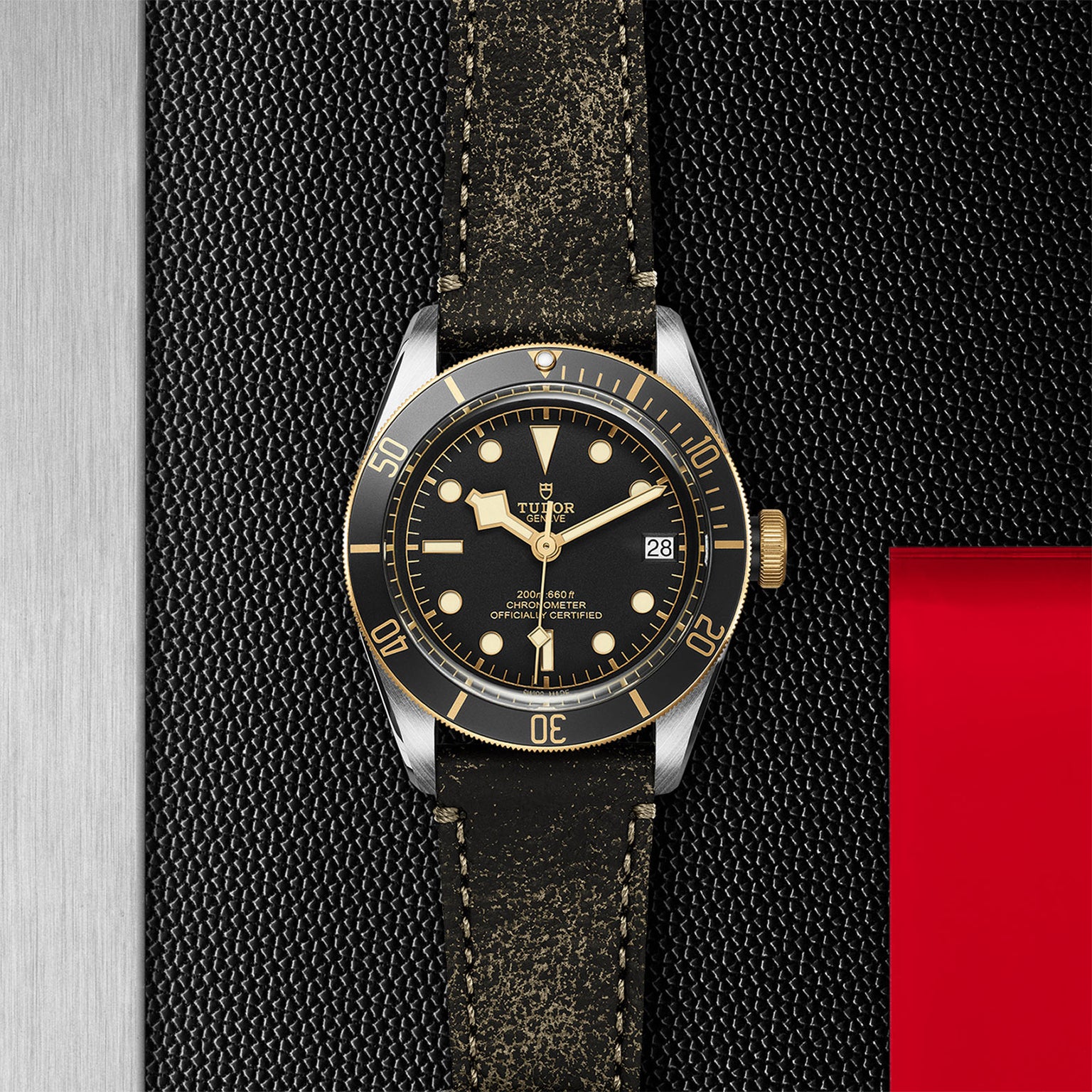 A TUDOR Black Bay S&G wristwatch with a black and gold bezel, black dial, gold accents, and a distressed black leather strap rests on a textured black and silver background—an icon among diving watches.