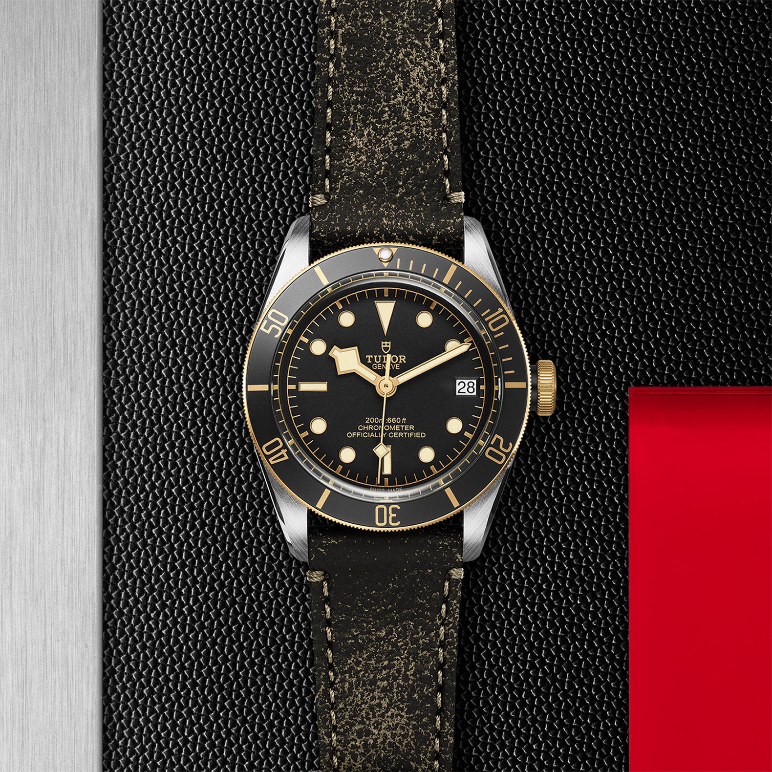 A TUDOR Black Bay S&G wristwatch with a black and gold bezel, black dial, gold accents, and a distressed black leather strap rests on a textured black and silver background—an icon among diving watches.
