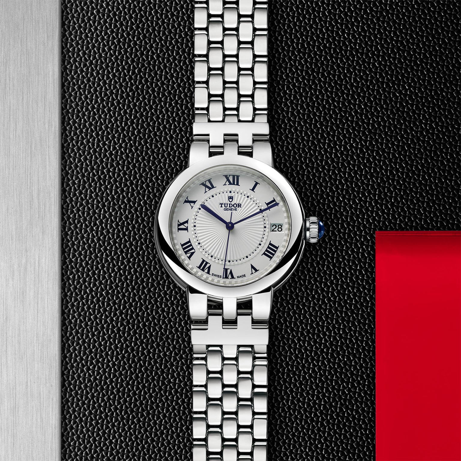 The TUDOR Clair de Rose wristwatch features a white dial, Roman numerals, and a metal link band, displayed on a textured black surface.