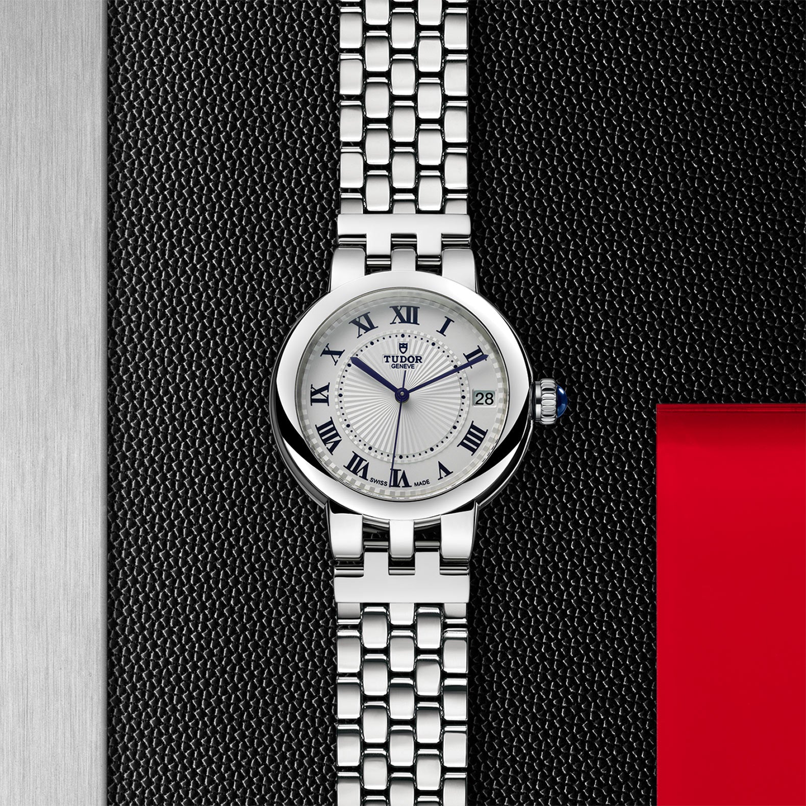 The TUDOR Clair de Rose wristwatch features a white dial, Roman numerals, and a metal link band, displayed on a textured black surface.