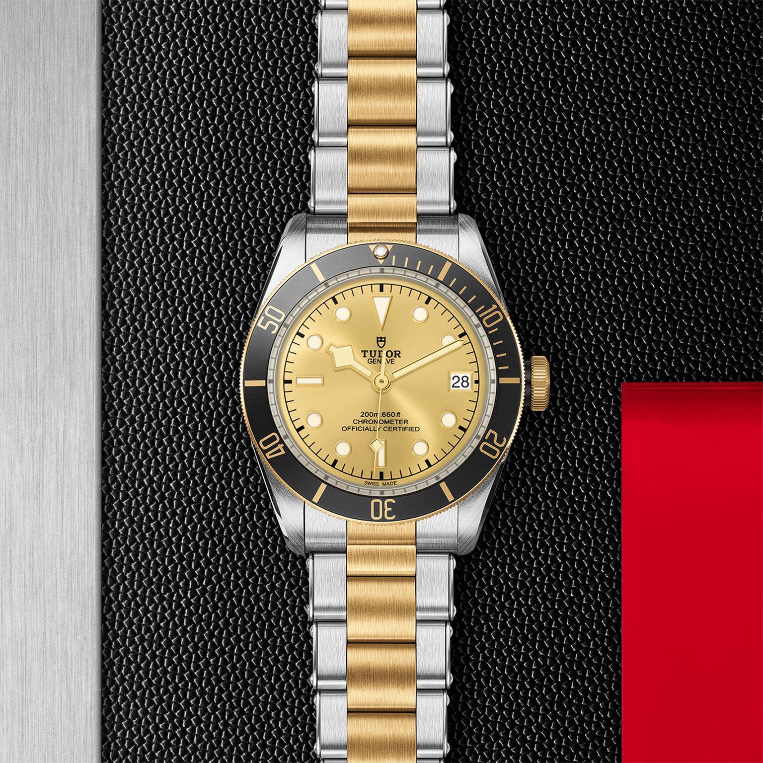 A two-tone TUDOR Black Bay S&G wristwatch with a gold dial and black bezel rests on a textured black surface beside a red accent. Its self-winding mechanical movement keeps the date window set to the 28th.
