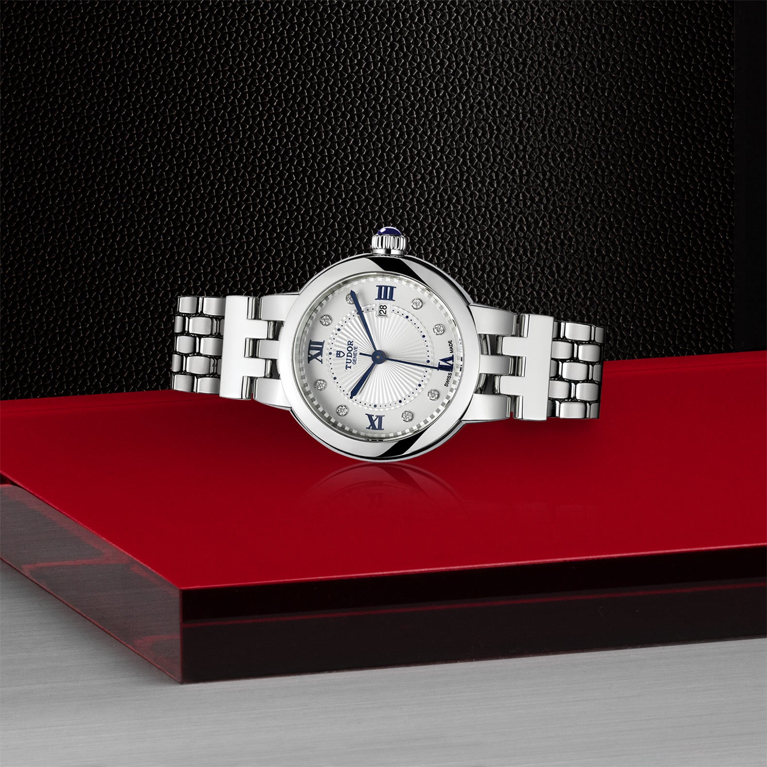 The TUDOR Clair de Rose, a silver luxury wristwatch with a white face, is displayed on a red and silver surface against a black background.
