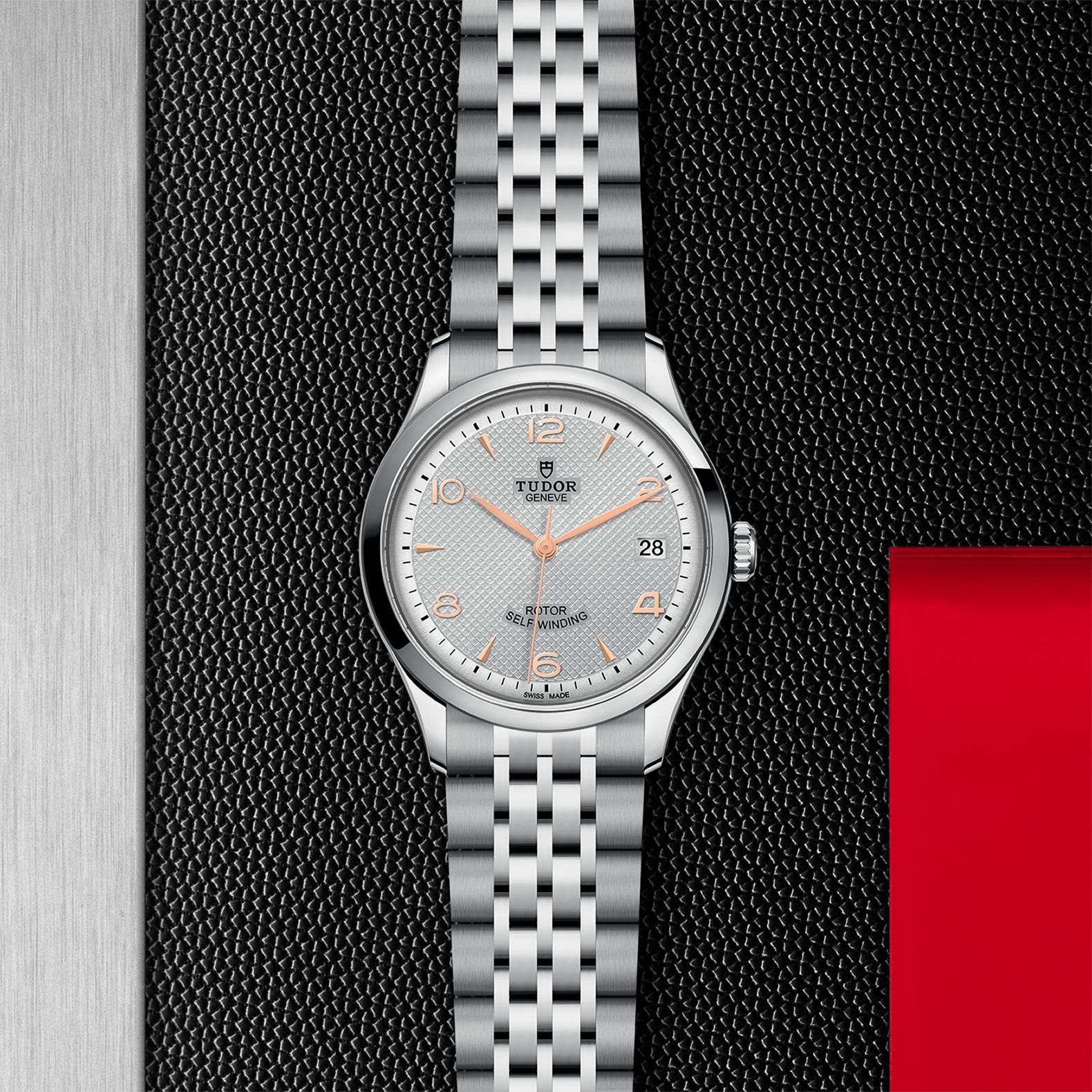 A TUDOR 1926 silver watch with orange-accented face and metal band is displayed on a textured black surface.
