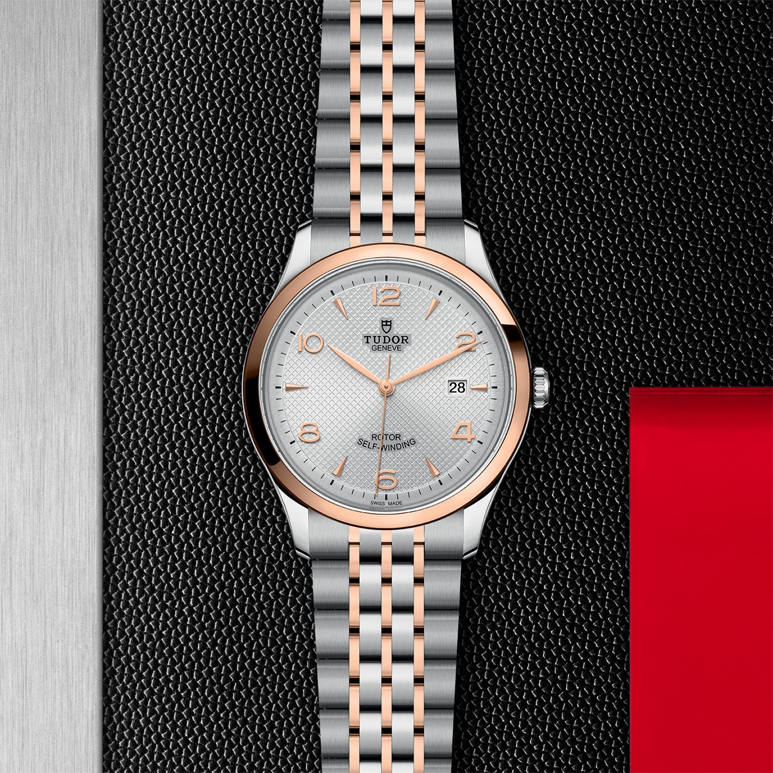 The TUDOR 1926 wristwatch, featuring a silver and rose gold metal band, silver dial, and date display, showcases timeless elegance against a textured black and metallic backdrop with a striking red accent.