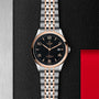 The TUDOR 1926 wristwatch, featuring stainless steel and rose gold with a black dial, gold hour markers, and date display, is displayed on a textured black and red background—an elegant mechanical timepiece for any collection.