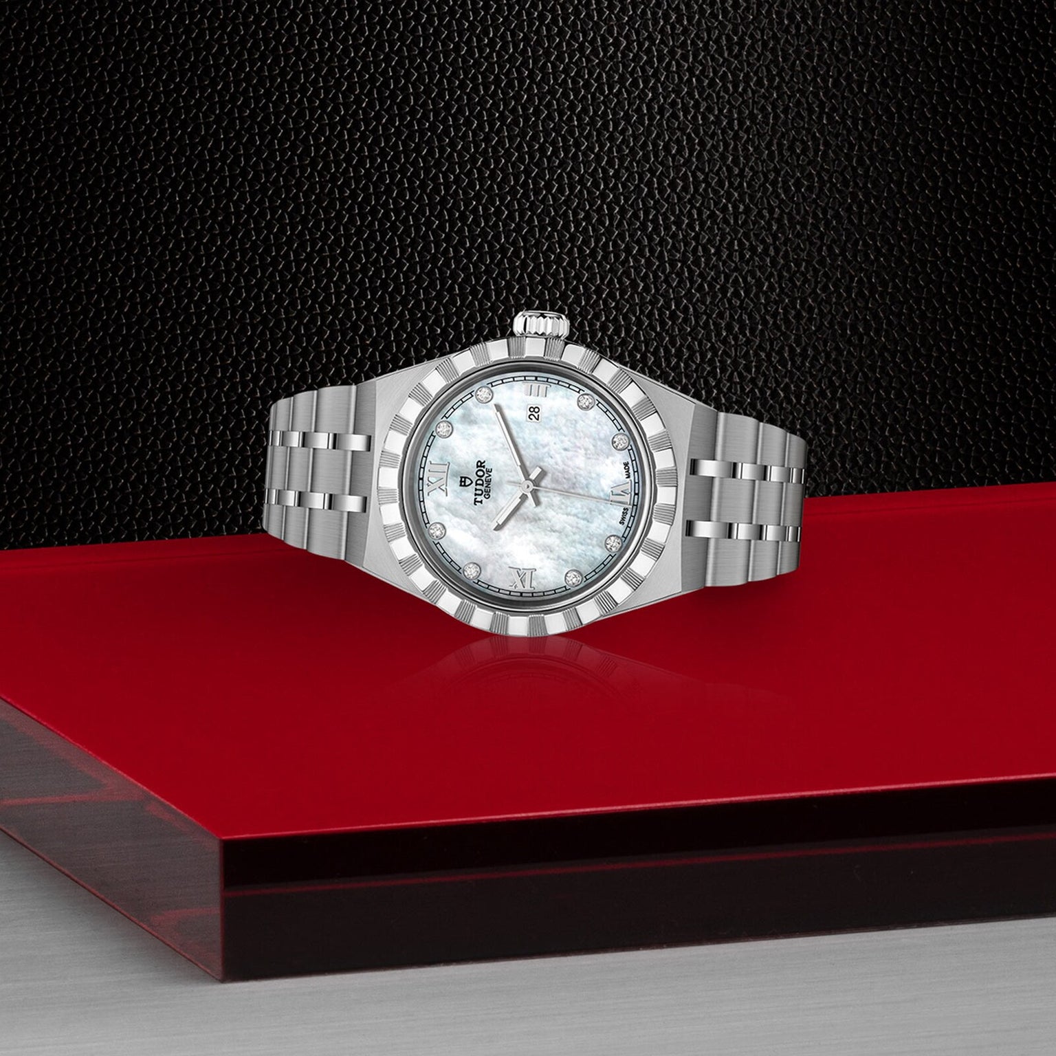 The TUDOR Royal, a silver wristwatch with a white dial and date display, is showcased on a glossy red and transparent platform, capturing the automatic sport-chic style against a textured black background.