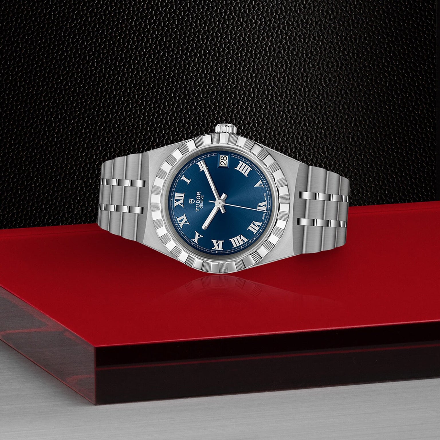 The TUDOR Royal sport-chic watch features a stainless steel bracelet, blue dial with Roman numerals, and date display, elegantly showcased on a striking red and black surface.
