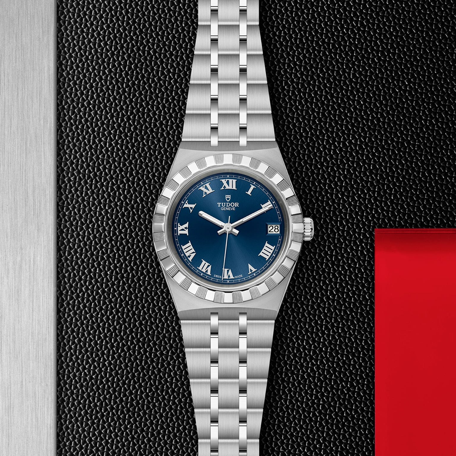 A silver TUDOR Royal wristwatch with a blue dial, Roman numerals, and date display lies on a textured black surface near red and silver panels, embodying the sport-chic style for which the TUDOR Royal is renowned.