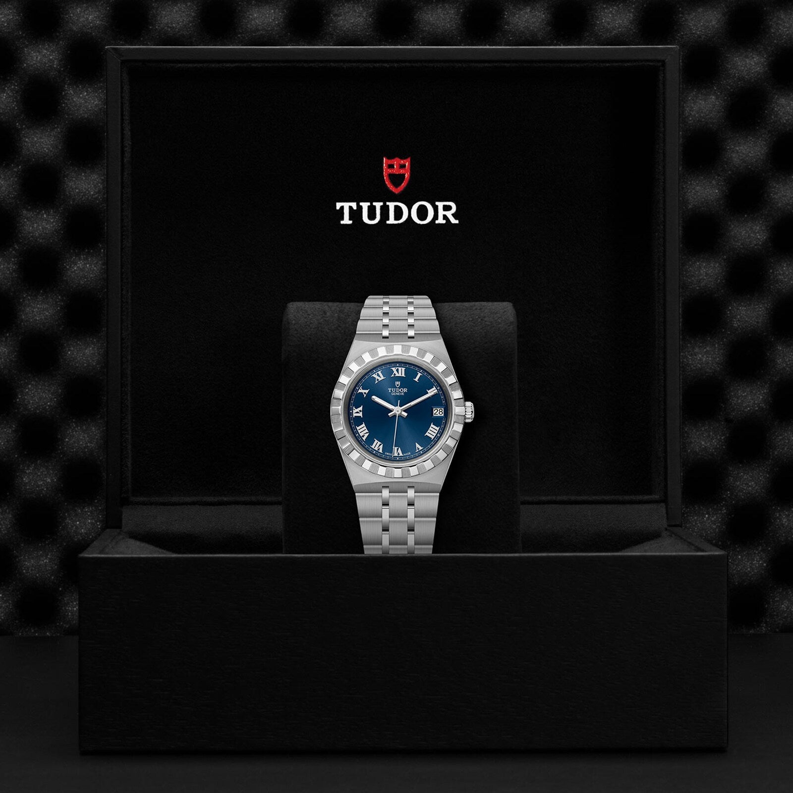 A TUDOR Royal watch with a blue dial and silver metal bracelet stands upright in a black Tudor-branded presentation box.
