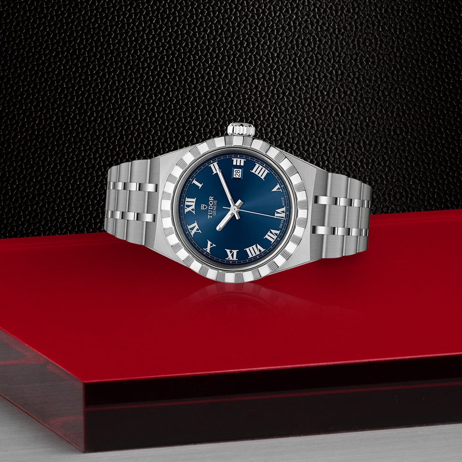 The TUDOR Royal, a silver wristwatch with a blue face and Roman numerals, is displayed on a red and black textured surface.
