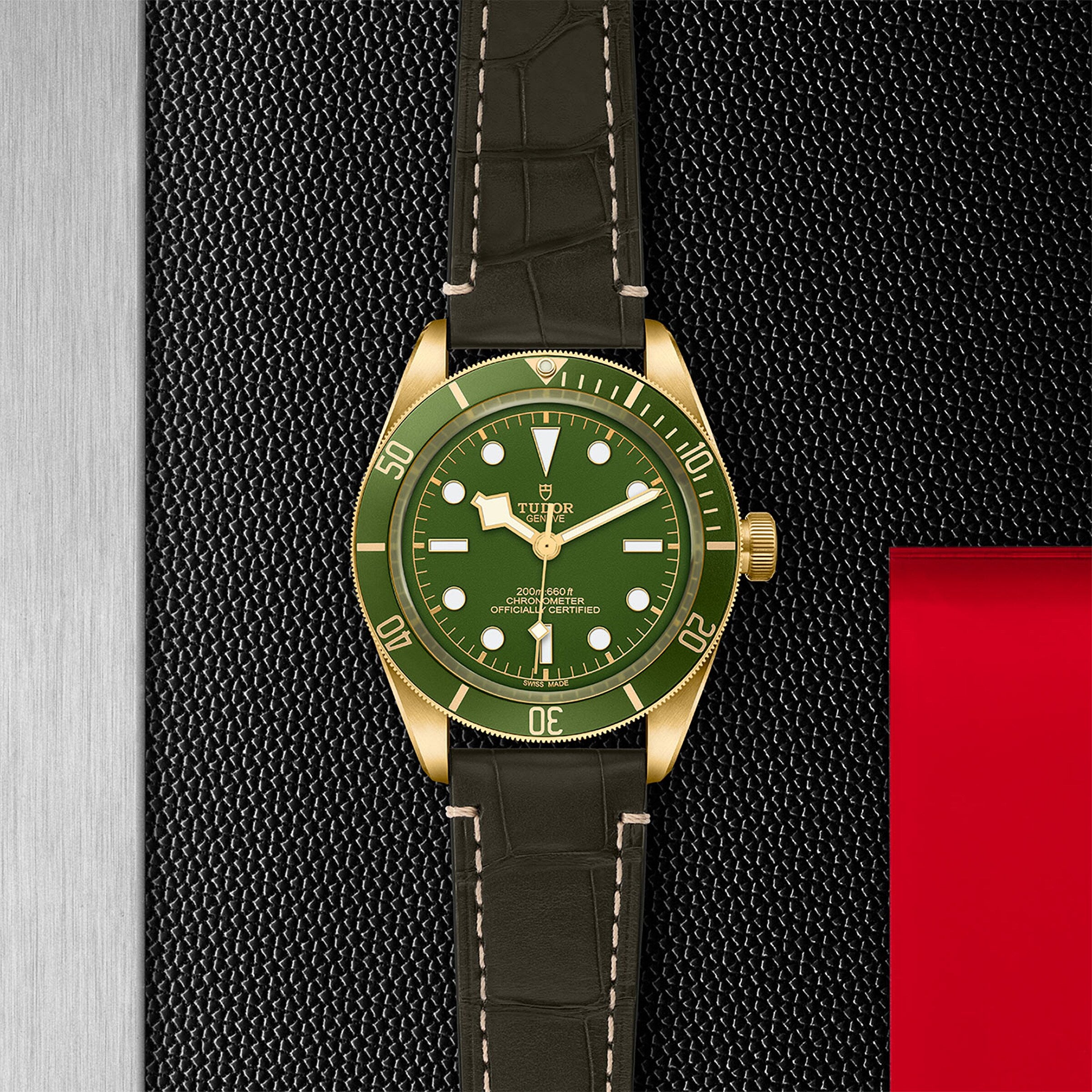 The TUDOR Black Bay 58 18K, featuring a green and gold design with a black leather strap, is displayed on a textured black surface accented with metal and red details.