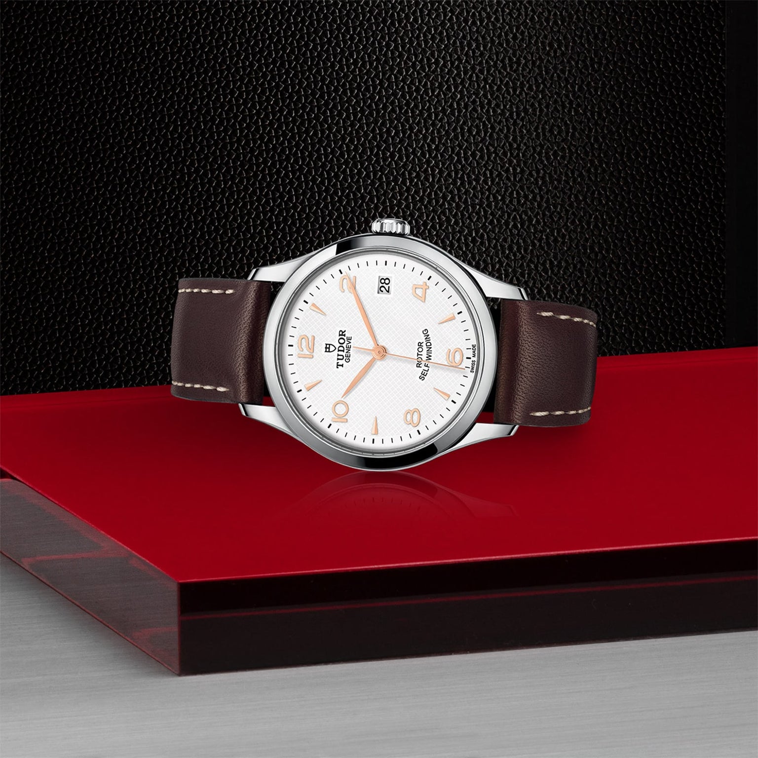 A TUDOR 1926 watch with a brown leather strap and white dial is showcased on a red platform against a textured black background, highlighting the classic elegance of this timepiece.