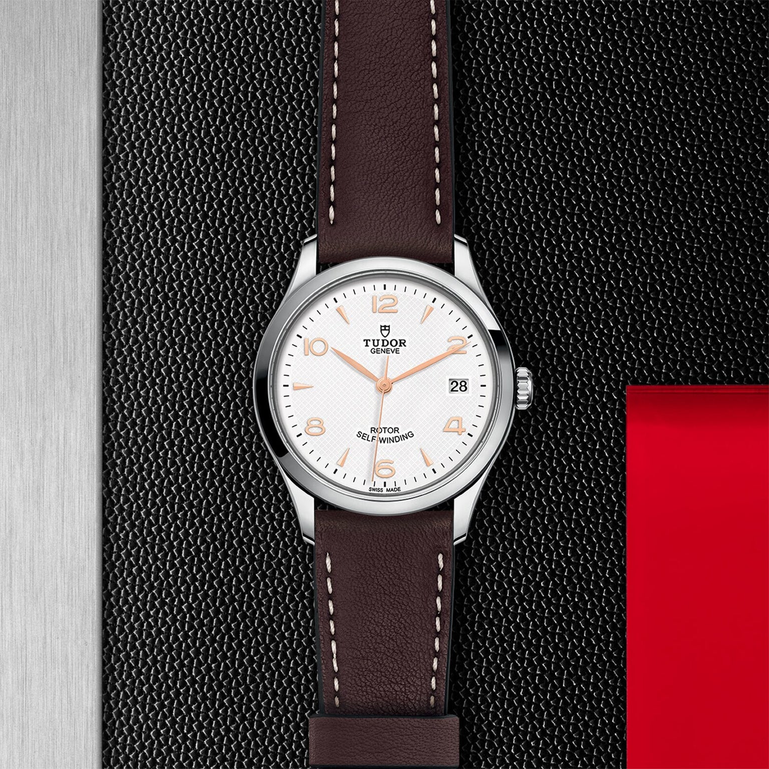 A TUDOR 1926 wristwatch featuring a white dial, brown leather strap, silver bezel, and date display rests on a textured black surface with red and metallic details—perfect for fans of classic elegance.