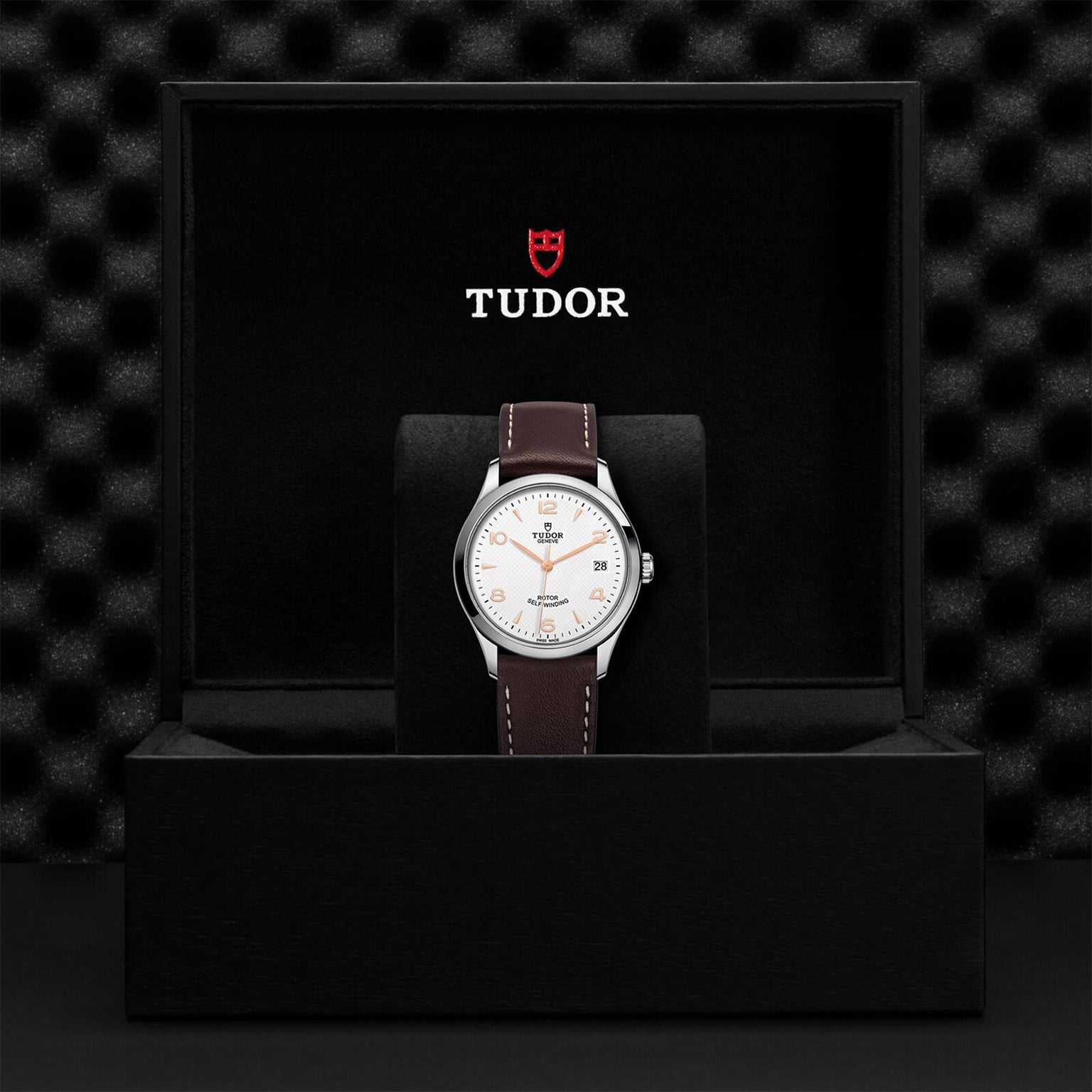 A TUDOR 1926 wristwatch with a brown leather strap and white dial is displayed in an open black box with the TUDOR logo on the inside lid, highlighting the classic elegance of this mechanical timepiece.