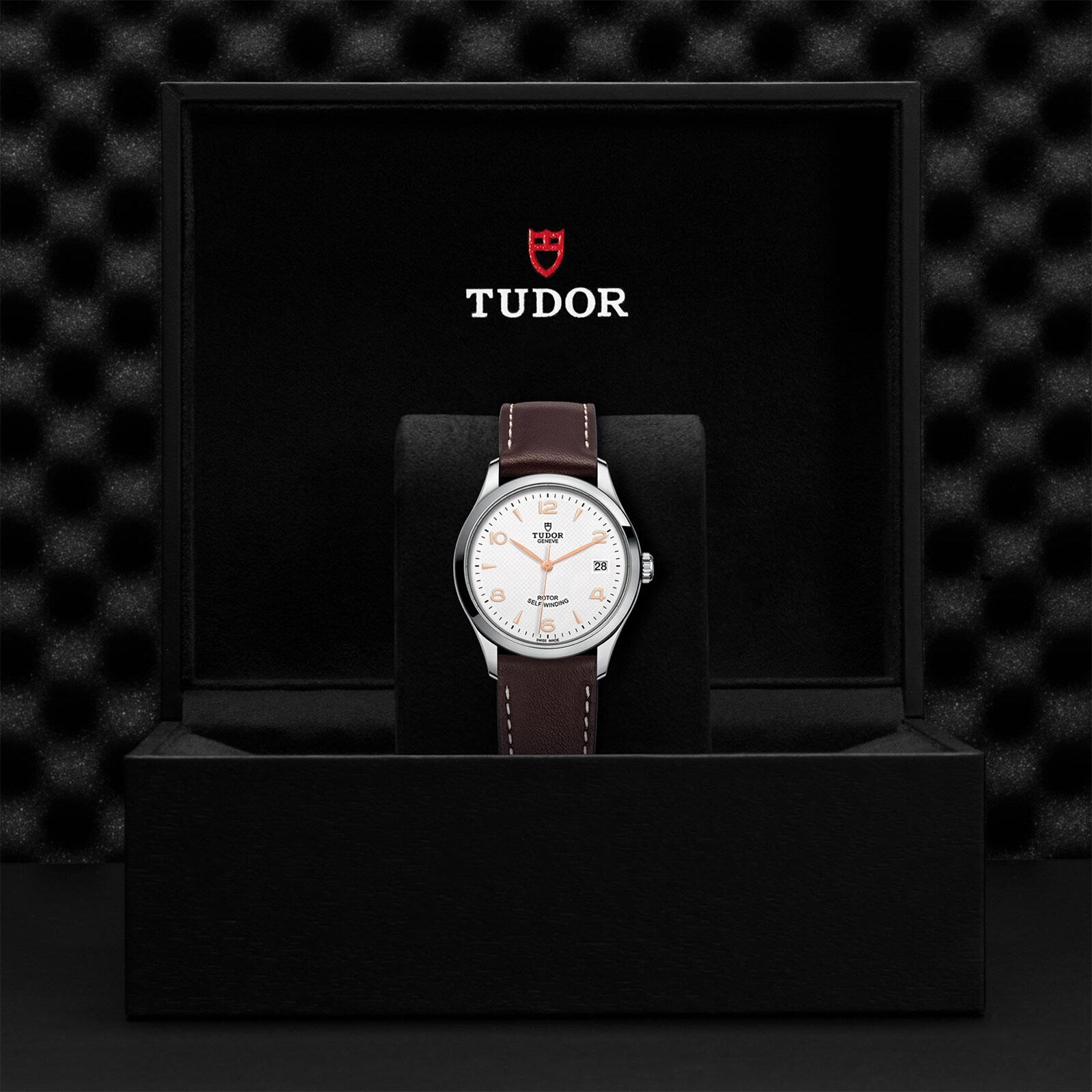 A TUDOR 1926 wristwatch with a brown leather strap and white dial is displayed in an open black box with the TUDOR logo on the inside lid, highlighting the classic elegance of this mechanical timepiece.