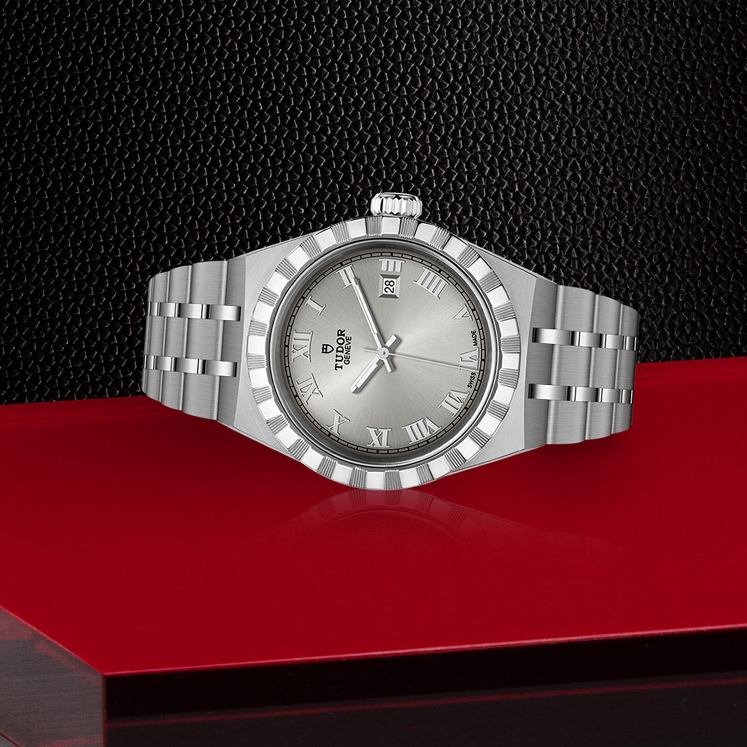 A TUDOR Royal wristwatch with a metallic band, Roman numerals, and date window is displayed on a red and black textured surface, embodying the sport-chic style of the TUDOR Royal collection.