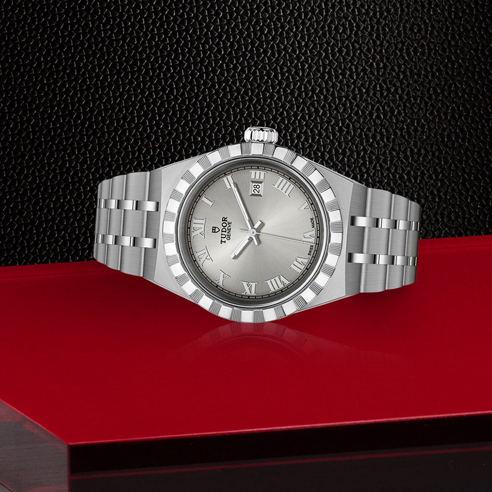 A TUDOR Royal wristwatch with a metallic band, Roman numerals, and date window is displayed on a red and black textured surface, embodying the sport-chic style of the TUDOR Royal collection.