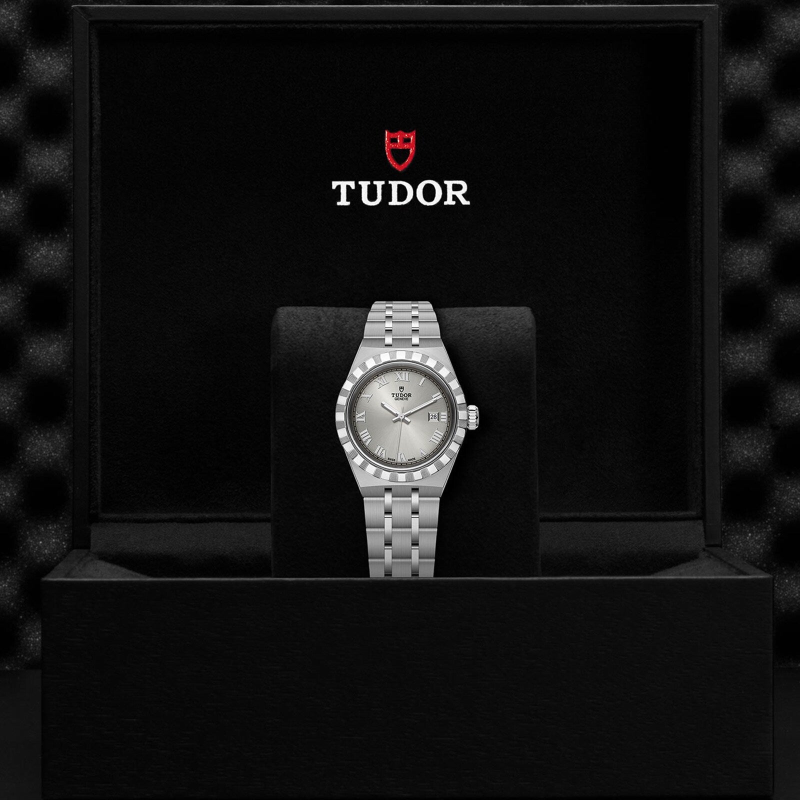 A silver TUDOR Royal wristwatch with a metal bracelet stands upright in a black presentation box, showcasing the TUDOR logo and name inside the lid—an elegant example of sport-chic timepieces.