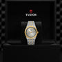 A TUDOR Royal wristwatch with a silver and gold integrated bracelet is presented in an open black box, featuring the TUDOR logo and name above the watch.