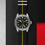The TUDOR Black Bay Pro features a black and yellow striped fabric strap, black dial, date display, and silver bezel—displayed on a textured black-metal surface with red accent—ideal for Black Bay fans.