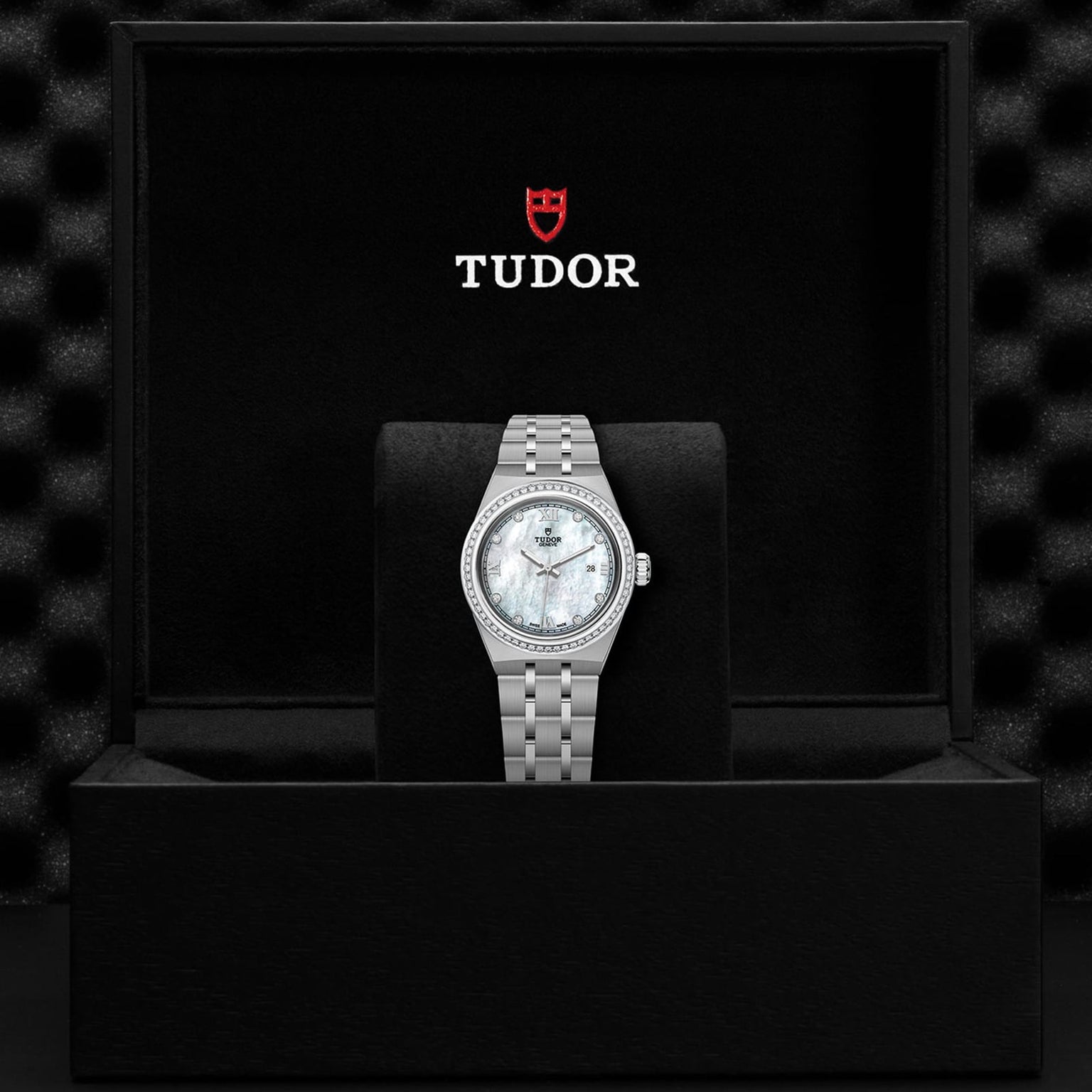 A TUDOR Royal wristwatch with a silver metal band and white face is displayed in an open black TUDOR-branded box.