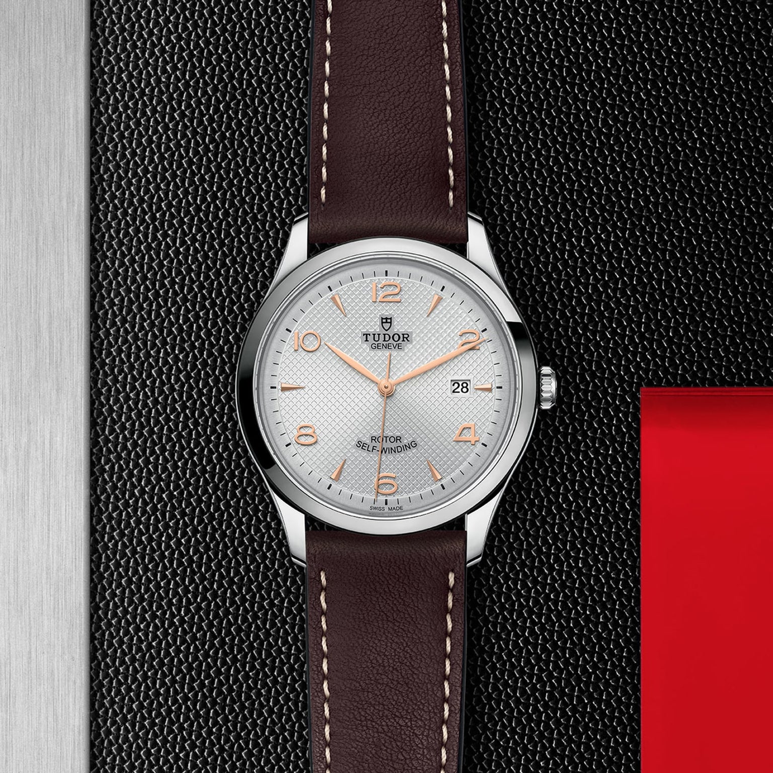 A TUDOR 1926 wristwatch featuring a white dial with orange accents, a brown leather strap, and date display rests on a textured black surface with red and metallic details, honoring the heritage of mechanical watches.
