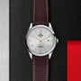 A TUDOR 1926 wristwatch featuring a white dial with orange accents, a brown leather strap, and date display rests on a textured black surface with red and metallic details, honoring the heritage of mechanical watches.