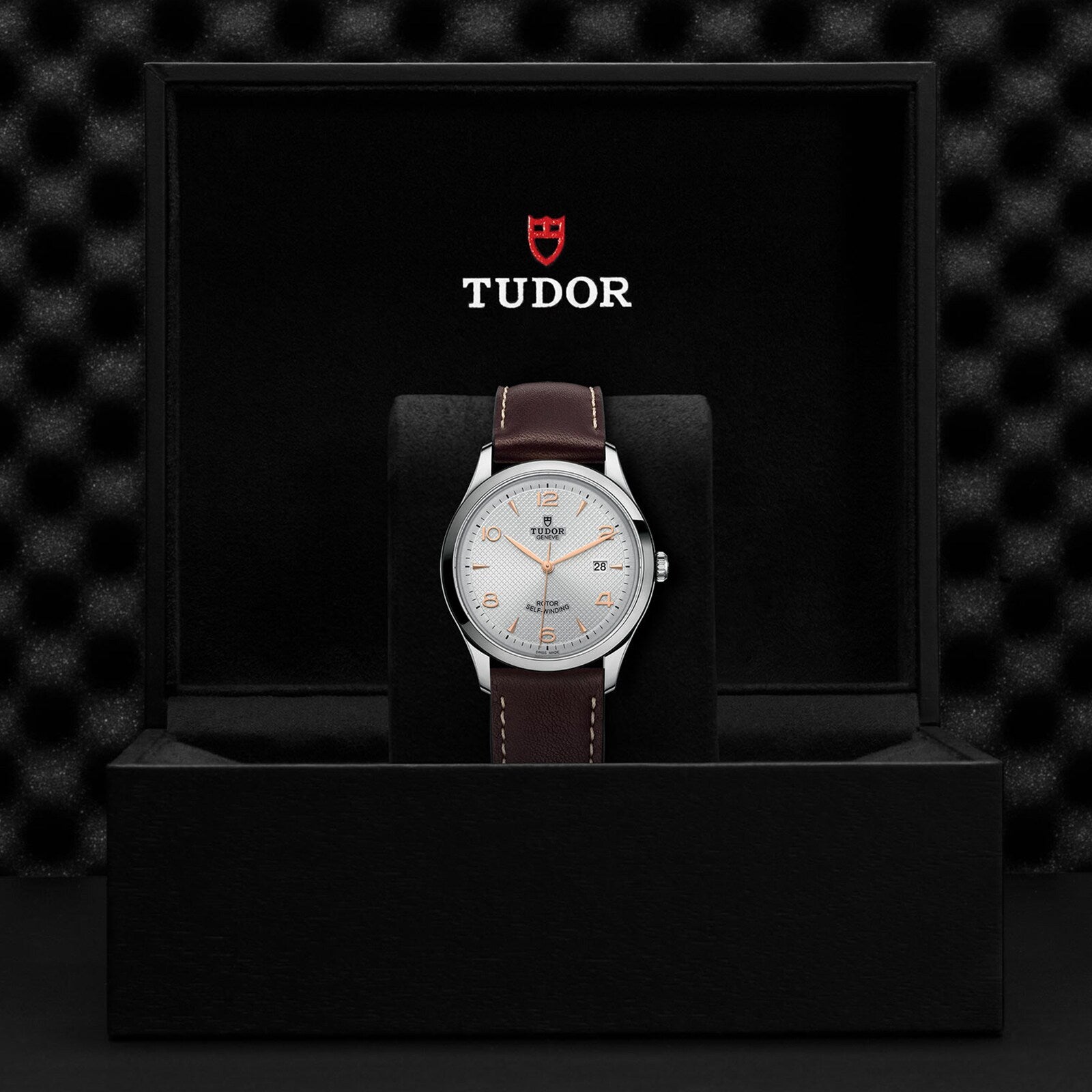 A TUDOR 1926 wristwatch with a brown leather strap and silver dial is displayed upright in a black presentation box with the TUDOR logo inside the lid.