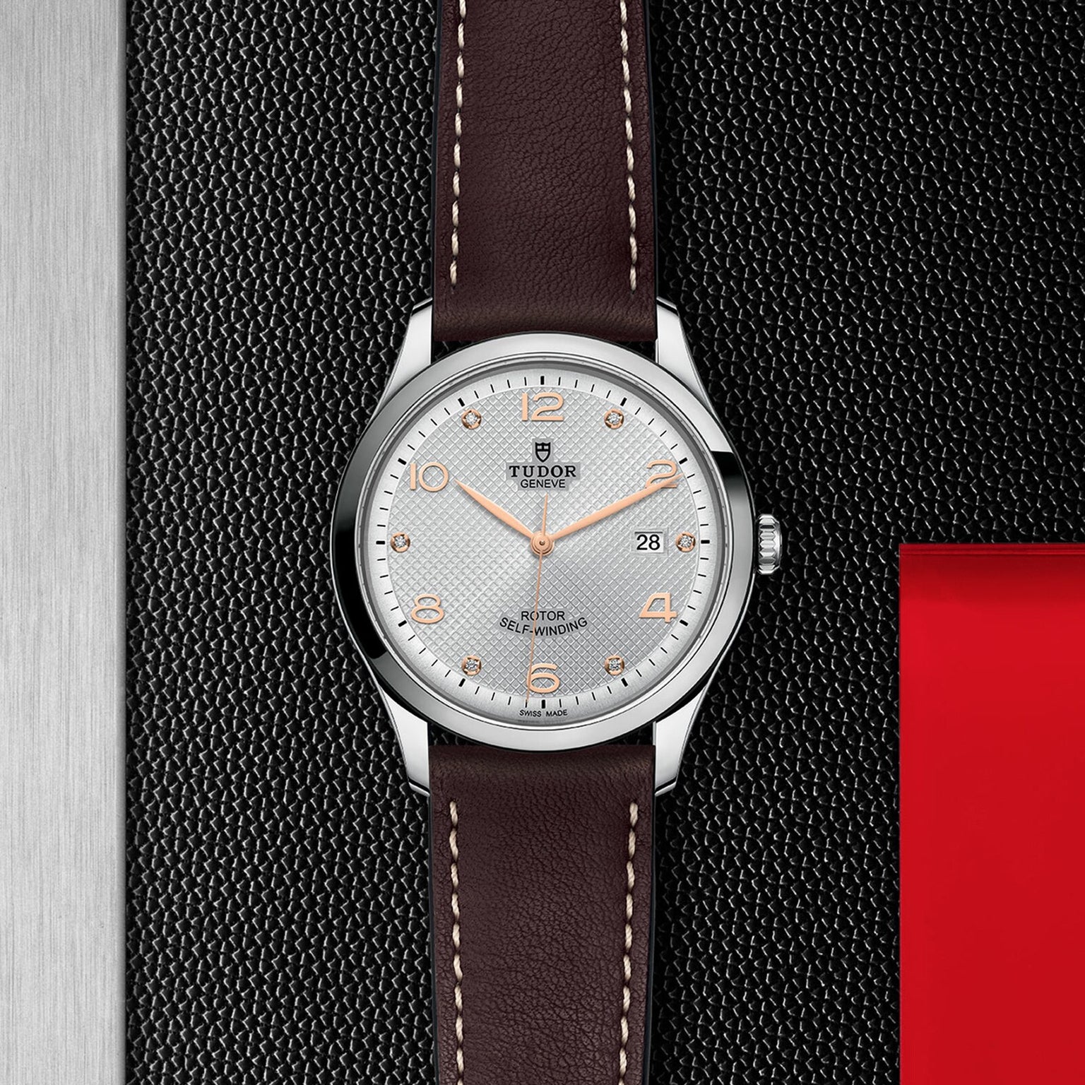 The TUDOR 1926 wristwatch features a white textured dial with orange numerals, date display, and a brown leather strap, elegantly presented on a textured black surface.