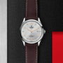 The TUDOR 1926 wristwatch features a white textured dial with orange numerals, date display, and a brown leather strap, elegantly presented on a textured black surface.