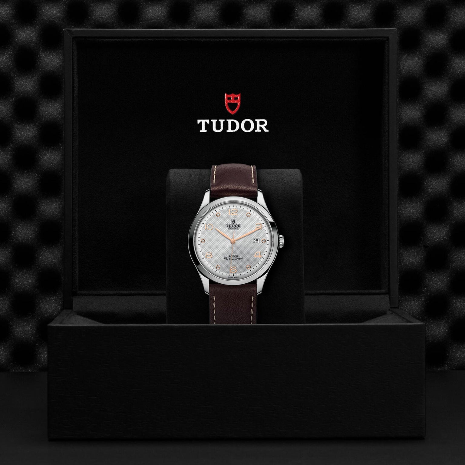 The TUDOR 1926 wristwatch, featuring a silver dial and brown leather strap, is displayed upright in a black box with the Tudor logo inside—a timeless choice for mechanical watch enthusiasts.