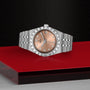 The TUDOR Royal is a silver wristwatch with a rose gold face and diamond markers, shown on a red and black background.